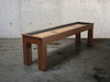 3/4 lifestyle view of the 9-foot Paula Parsons Shuffleboard Table by Doc & Holliday in Grey Walnut finish, featuring solid acacia wood, recessed scoring, and a maple butcher block playfield with modern Parsons-style legs, displayed against a sleek industrial backdrop.