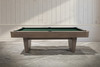 A full side profile of the Doc & Holliday Preakness slate pool table, photographed in a quiet architectural setting to emphasize its low, grounded stance. The Glazed Pearl maple finish reads soft and refined against the neutral backdrop, while the Championship green felt adds a classic contrast that keeps the table rooted in traditional play.