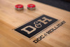 Close-up of the Savant shuffleboard table playfield by Doc & Holliday showing the branded maple surface with red shuffleboard pucks, emphasizing gameplay detail and surface finish.