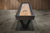 End view of the Savant shuffleboard table by Doc & Holliday highlighting the maple butcher block playfield with inlaid Doc & Holliday logo, recessed scoring system along the rail, and bold industrial steel V leg base.
