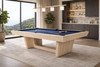 Doc & Holliday Kaci 8-ft slate pool table in Natural Ash finish with navy blue felt, photographed in a modern luxury game room with floor-to-ceiling windows, light oak wood grain detail, contemporary pedestal base, and interior design-inspired setting.