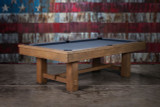 ¾ hero view of The Hank Pool Table by Doc & Holliday in Oaklee finish, photographed with a USA flag backdrop. Professionally staged, showing solid oak frame, finger-jointed rails, and slate playfield in a rustic Americana setting.
