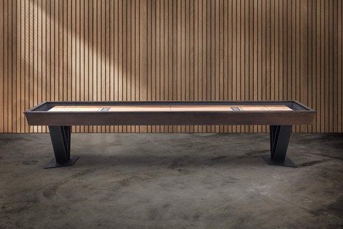 Savant shuffleboard table by Doc & Holliday in Brown Sugar finish, photographed in a modern studio with wood slat walls and concrete flooring, showcasing the table’s long, clean profile, industrial steel V legs, and maple shuffleboard playfield.