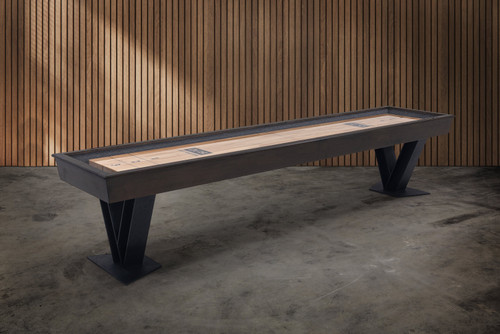 Angled lifestyle view of the Savant shuffleboard table by Doc & Holliday, featuring a Brown Sugar wood cabinet, steel V-shaped legs, recessed gutters, and a precision maple playing surface designed for modern game rooms.