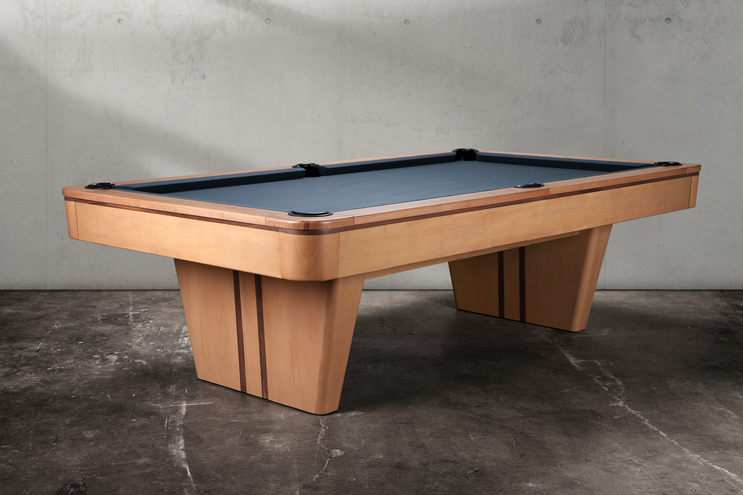 The Doc & Holliday Preakness Slate Pool Table shown in a 3/4 studio angle with a natural maple finish and deep blue felt. Its bold pedestal-style legs with walnut inlay accents give it a mid-century modern stance, while the three-piece slate playfield ensures precision and true roll. A striking Americana-inspired slate billiard table designed to anchor both contemporary and classic spaces.