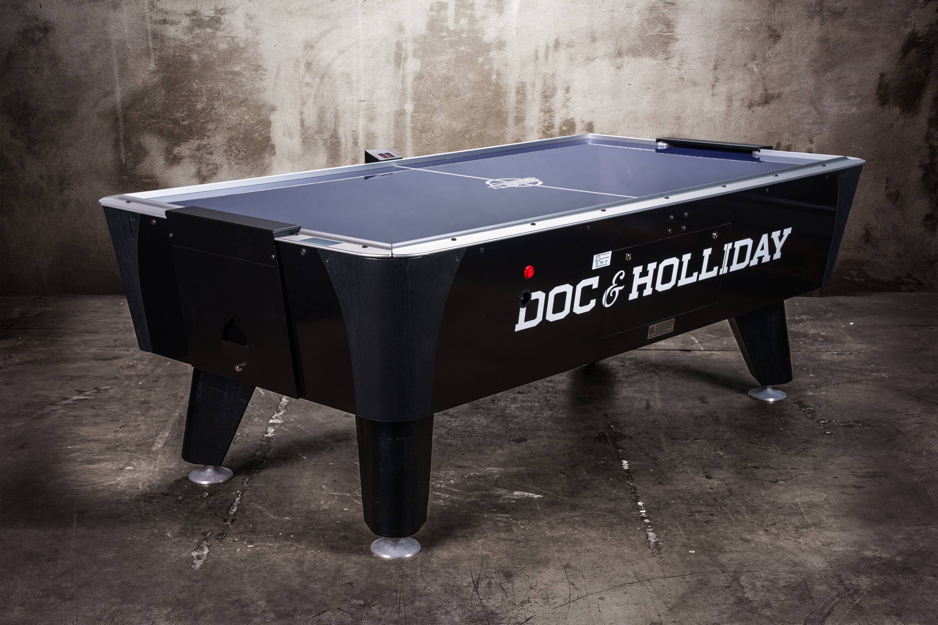 Angled 3/4 view of the Doc & Holliday Air Hockey Table during a photoshoot, capturing the sleek cabinet edges, tapered legs with leveler feet, and electronic scoring unit. The dramatic lighting emphasizes the craftsmanship and durability, positioning the table as both furniture and entertainment centerpiece.
