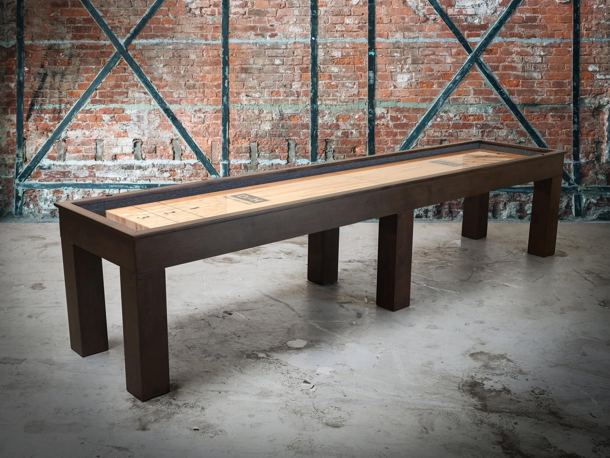 Paula Parsons Shuffleboard Table by Doc & Holliday in Antique Coffee finish styled in a modern game room with brick walls and concrete flooring, showing its clean Parsons frame and warm alder wood tones.