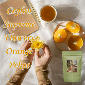 Hands hold a yellow flower next to a notebook, a cup of tea, and a plate of orange slices on a white fabric background.