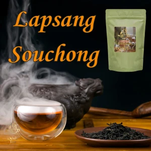 A small glass cup of steaming amber tea rests beside a brown dish of loose smoked black tea leaves on a wooden table.