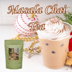 A clear plastic cup holds iced masala chai topped with whipped cream and spice, alongside holiday ornaments on a white surface.