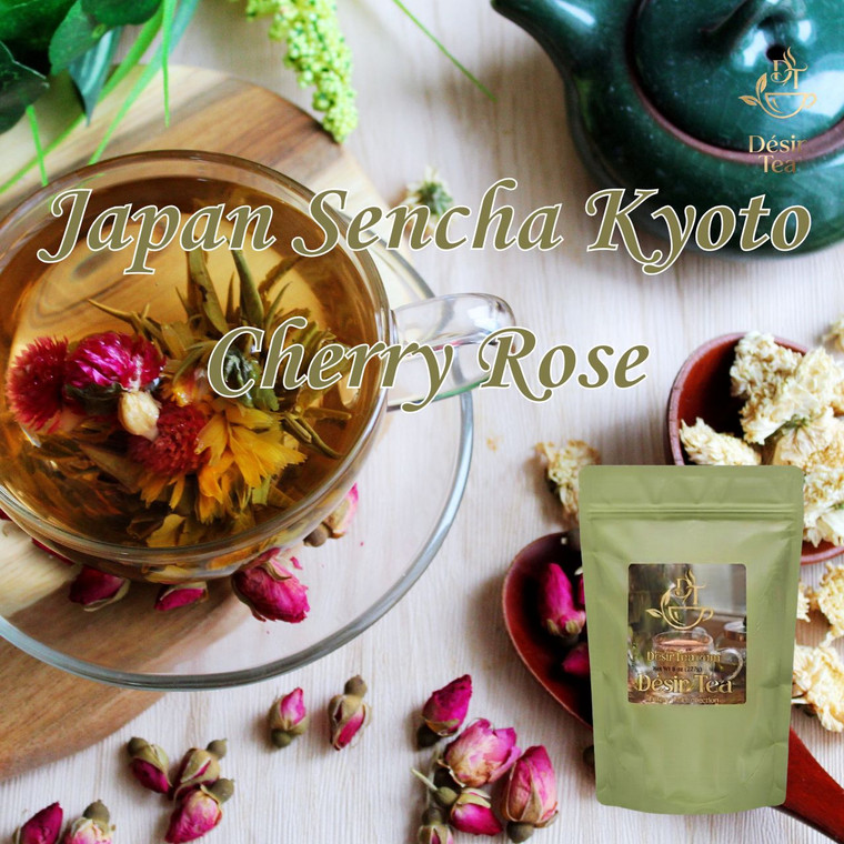 A transparent glass cup holds a blooming tea flower with red and yellow petals next to a Desir Tea pouch and scattered rosebuds.