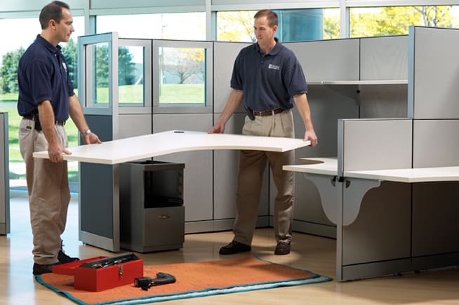 Two technicians positioning an L shaped desk during office cubicle installation.
