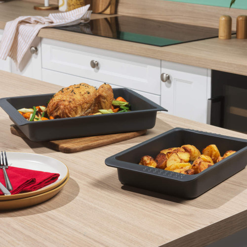 Two grey roasting trays with roast chicken and potatoes on a kitchen worktop.