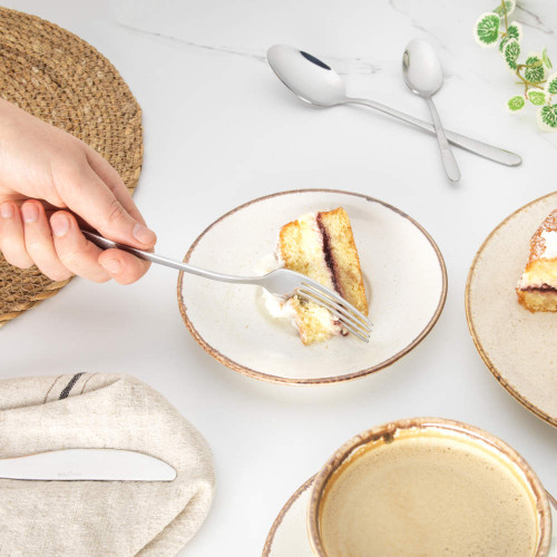 Hand holding fork with cake slice and cream on ceramic plate, coffee and cutlery nearby.