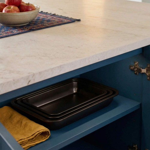 Set of three black roasting trays on blue kitchen shelf, shown with folded yellow tea towel.