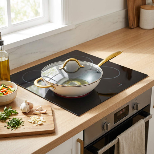 Cream non-stick sauté pan with gold handles and lid on induction hob in modern kitchen