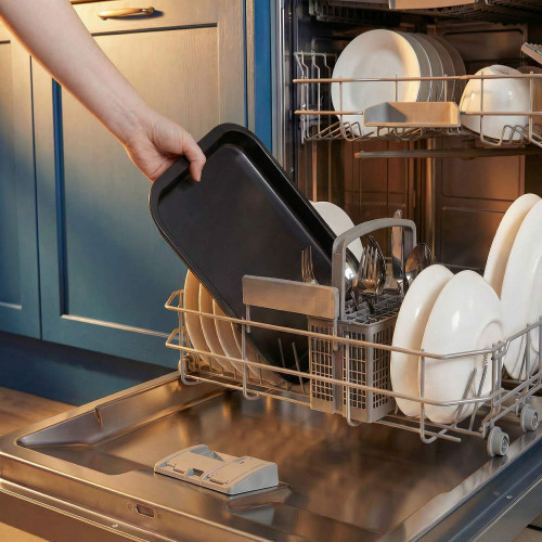 Hand placing black oven tray in dishwasher with clean plates and cutlery