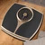 Mechanical bathroom scale in black and gold, shown on wood floor, top view.