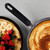 Non-stick frying pan with cooked pancake, next to filled crepe topped with fresh berries and banana.