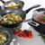 Set of non-stick black pans with cooked food and fresh tomatoes on white surface