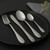 Four-piece stainless steel cutlery set on black plate, includes fork, knife, dessert spoon and teaspoon.