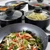 Set of non-stick black saucepans and frying pans with glass lids, shown with cooked food.