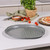 Grey non-stick pizza tray with holes, shown on kitchen counter near fresh tomatoes and herbs
