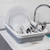 Grey and white collapsible dish drainer with clean plates, mugs, and cutlery beside sink