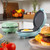 Blue electric breakfast maker shown open with fried egg, burger and tomatoes in kitchen.