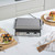 Stainless steel contact grill on kitchen counter, lid closed, with sandwiches and kebabs nearby
