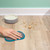 Hand wiping pet crumbs from wooden floor near dog bowl and pink pet bed