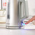 Silver electric kettle with visible water gauge and blue power switch being pressed