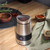 Salter stainless steel electric grinder on wooden countertop, surrounded by herbs and spices