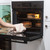 Person removing round cake tin from oven using tea towel, other tins inside