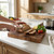 Person slicing rustic bread on wooden chopping board with lemon, tomatoes and basil nearby