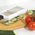 White mandoline slicer with black handle slicing cucumber on a chopping board with tomatoes