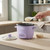Purple electric rice cooker on kitchen worktop, rice being poured in, lid and bowl beside.