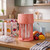Pink blender with clear jug on kitchen counter, surrounded by juice cartons and fresh fruit.