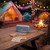 Blue portable Bluetooth speaker on picnic table at campsite with tents and campfire at dusk