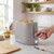 Grey 2-slice toaster with bread, hand pressing lever, kitchen counter setting