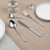 Four silver and light blue cutlery pieces on beige tablecloth, shown with plates and glasses