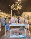 Retail boutique space showcasing the ShopWildThings Roses & Greenery Hanging Canopy, a round floral chandelier measuring 46 inches wide by 16 inches long, suspended over a display table with folded apparel and accessories. The chandelier features artificial roses and greenery in soft pink tones, adding a dramatic, elegant element to the boutique’s visual merchandising strategy.