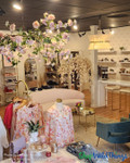 Chic women’s boutique decorated with the ShopWildThings Adalee Cherry Blossom Tree in blush pink and a round floral chandelier canopy of artificial roses and greenery. The tree’s full bloom branches create a romantic atmosphere near a soft blush velvet couch, while the floral chandelier provides overhead interest above a curated display of spring apparel, accessories, and gift items. Ideal for boutique decor and seasonal visual merchandising.