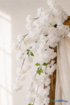 41" White Cascading "Spring Blossom" Spray – Extra-Full Silk Floral Waterfall from ShopWildThings.com styled on a wooden wedding arch with soft white draping, shown in a close-up detail view with crisp white blooms and trailing floral sprays.