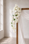 41" Cream White Cascading "Spring Blossom" Spray – Extra-Full Silk Floral Waterfall from ShopWildThings.com attached to a simple wooden wedding arch in natural window light, with white fabric draping and cream white floral stems cascading downward.