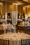 Luxurious wedding reception with round tables covered in white linens, each decorated with tall 38-inch Juliet crystal centerpiece risers featuring cascading crystal bead strands, surrounded by chiavari chairs and sparkling under elegant chandelier lighting.