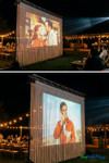 Wedding or outdoor dinner event featuring a classic movie projected onto a tall white string curtain backdrop from ShopWildThings, illuminated by warm market lights and supported by a truss frame.
