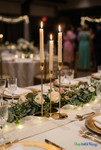 Gold long stem taper candle holder centerpiece with lit candles on elegant wedding reception table with greenery runner