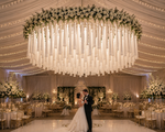 An elegant indoor wedding installation showcasing a custom white tassel chandelier suspended high above the couple’s first dance. The chandelier features layered oversized tassels hanging from a floral-covered circular frame with soft greenery accents. The elevated design creates an airy, luxurious look while allowing the dramatic tassels to frame the dance floor without overwhelming the space. Twinkling ceiling lights and romantic floral arrangements complete the upscale event setting.