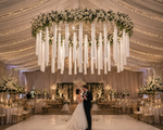 A dramatic statement chandelier installation featuring a dense canopy of oversized white string tassels suspended from a grand floral ring overflowing with white roses and greenery. The fuller layered tassel design creates incredible texture, movement, and visual depth while forming a luxurious floating centerpiece above the wedding dance floor. Soft warm lighting illuminates the thousands of flowing strands, creating an immersive high-end wedding environment perfect for luxury receptions, galas, and large-scale event décor.